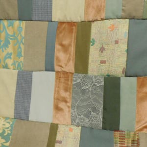 Könnte beinhalten: Eine Patchwork-Decke aus vertikalen Stoffstreifen in verschiedenen Texturen und Farben, darunter Salbeigrün, kupferfarbener Samt, Creme, Grau sowie gemusterte Paneele mit Blumen-, Spitzen- und geometrischen Designs, die rechteckig vernäht sind.
