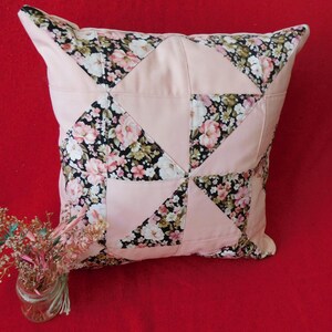 May include: A square throw pillow featuring a patchwork design of solid light pink fabric triangles alternating with a black fabric patterned with pink and white floral blossoms. A small glass jar containing dried flowers sits nearby.