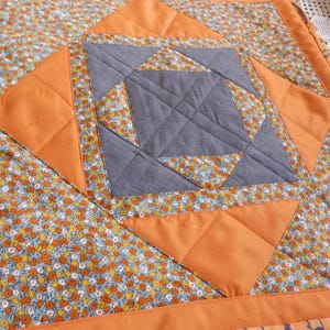 Könnte beinhalten: Ein quadratischer Patchwork-Quilt mit einem Blumenmuster in Orange, Gelb und Blau. Die Mitte zeigt ein graues geometrisches Design, umgeben von orangefarbenen Diamanten und einem durchgehenden orangefarbenen Rand. Der Quilt ist wahrscheinlich handgefertigt.