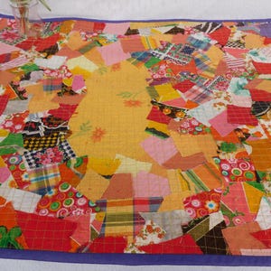 Könnte beinhalten: Ein rechteckiger Quilt mit einem chaotischen Patchwork aus verschiedenen Stoffresten in Rot-, Orange-, Gelb-, Rosa- und Grüntönen. Der Quilt ist mit einer einfarbigen violetten Stoffeinfassung versehen und zeigt ein großes zentrales gelbes Stofffeld.