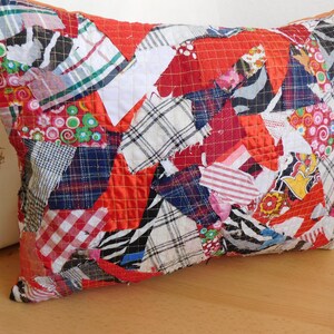 Könnte beinhalten: Ein rechteckiges Patchwork-Kissen mit einer lebendigen Mischung aus Stoffen in Rot, Weiß, Blau und gemusterten Designs. Das Kissen hat eine gesteppte Textur und eine warme Farbpalette, perfekt, um jedem Raum eine gemütliche Note zu verleihen.