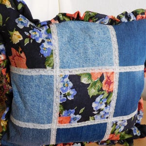 Könnte beinhalten: Quadratisches Zierkissen mit Patchwork-Design. Das Kissen hat Jeansquadrate, florale Stoffquadrate und weiße Spitzentrimmung. Die Kanten sind mit einem gerüschten floralen Stoff in Blau-, Orange- und Schwarztönen verziert.