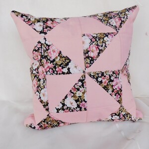 May include: A square throw pillow featuring a patchwork design of solid light pink fabric and a black fabric with a floral pattern of pink, white, and green flowers. The pattern is arranged in a pinwheel style with alternating triangular sections.