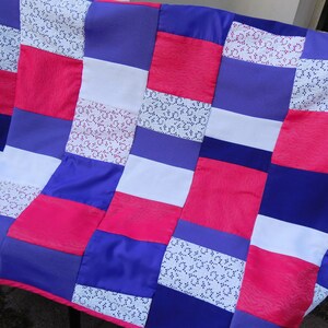 Könnte beinhalten: Ein Patchwork-Quilt mit einer lebendigen Farbmischung. Der Quilt zeigt ein Muster aus abwechselnden Quadraten und Rechtecken in Rosa-, Lila-, Weiß- und gemusterten weißen Stofftönen. Der Quilt ist aus einem seidigen Material gefertigt.
