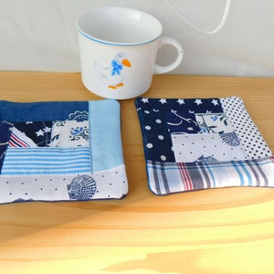 Könnte beinhalten: Zwei blaue und weiße Patchwork-Stoffuntersetzer. Die Untersetzer bestehen aus verschiedenen Stoffen, die zu einer quadratischen Form zusammengenäht sind. Die Untersetzer befinden sich auf einer Holzoberfläche.