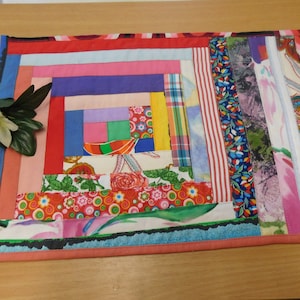 Könnte beinhalten: Ein farbenfrohes, rechteckiges Patchwork-Quilt mit einem Blockhaus-Design. Das Quilt weist eine Vielzahl von Stoffen in Rot, Rosa, Blau und floralen Mustern auf. Die Kanten sind mit einem rosa Rand versehen. Eine weiße Blume und grüne Blätter befinden sich links.