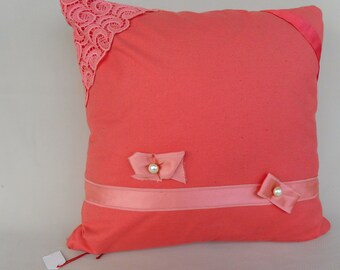 May include: Coral-colored decorative pillow with pink lace corner detail, a pink ribbon, and two small bows with pearl accents. The pillow is square-shaped and has a soft, textured appearance, suitable for home decor.