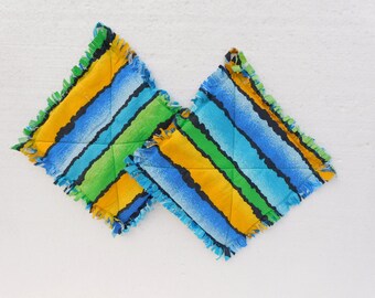May include: Two square, quilted pot holders with a colorful striped pattern. The design features blue, green, yellow, and black stripes. The edges are trimmed with a ruffled, fringed border. The pot holders are on a white background.
