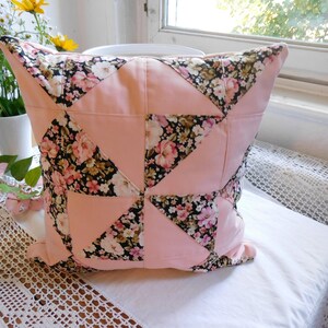 May include: A square throw pillow featuring a patchwork design of solid light pink fabric triangles alternating with a black fabric patterned with pink, white, and yellow floral blossoms. The pillow has a visible seam construction throughout.