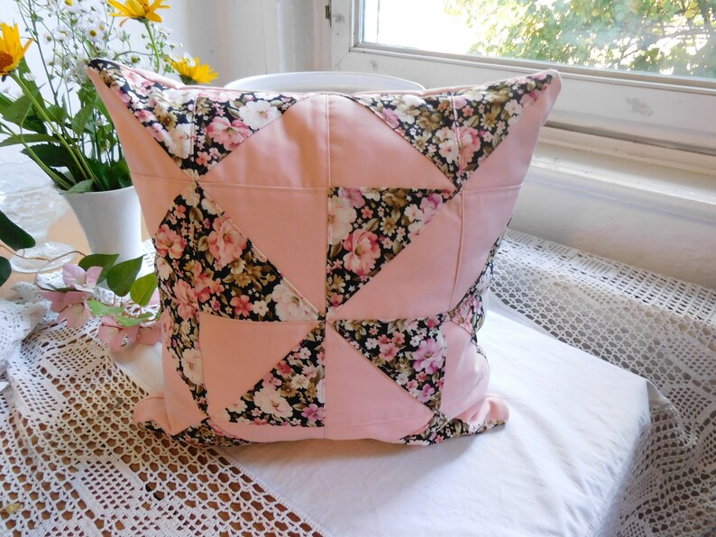 May include: A square throw pillow featuring a patchwork design of solid light pink fabric triangles alternating with a black fabric patterned with pink, white, and yellow floral blossoms. The pillow has a visible seam construction throughout.