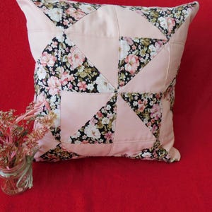 May include: A square throw pillow featuring a patchwork design of solid light pink fabric and a black floral print fabric with pink, white, and green flowers. The pillow is constructed with triangular and square panels arranged in a geometric pattern.