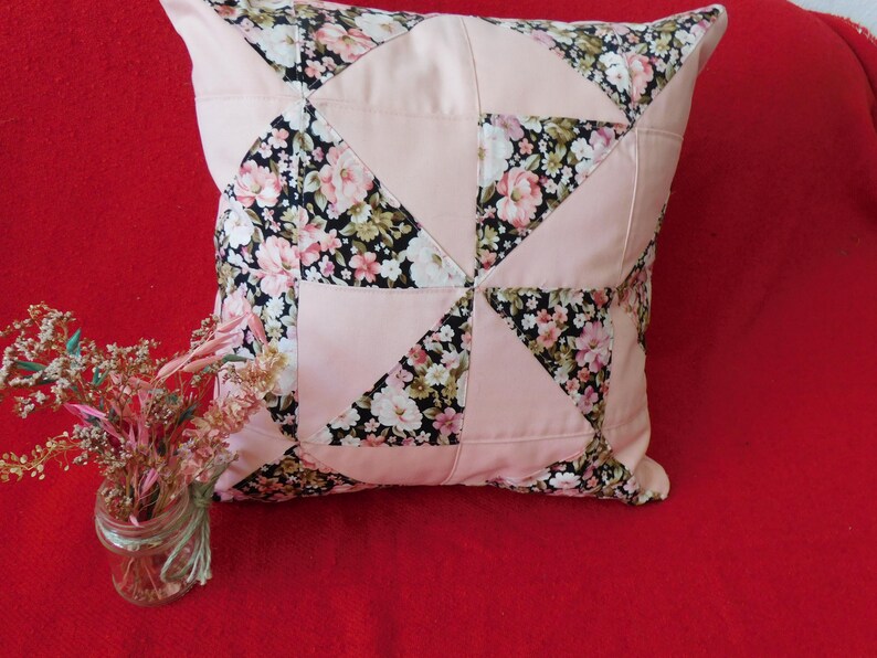 May include: A square throw pillow featuring a patchwork design of solid light pink fabric and a black floral print fabric with pink, white, and green flowers. The pillow is constructed with triangular and square panels arranged in a geometric pattern.