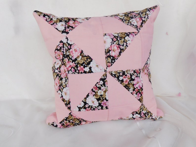 May include: A square throw pillow featuring a patchwork design of solid light pink fabric and a black fabric with a floral pattern of pink, white, and green flowers. The pattern is arranged in a pinwheel style with alternating triangular sections.