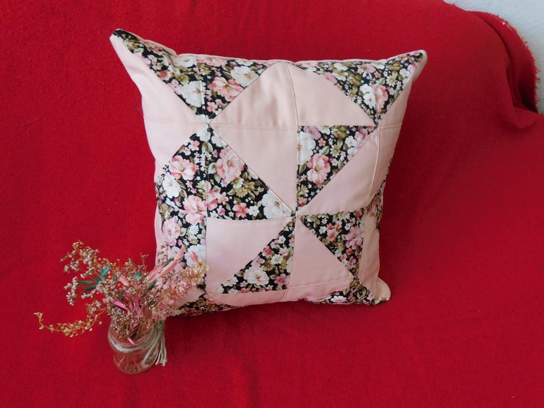 May include: A square throw pillow featuring a patchwork design of solid light pink fabric triangles alternating with a black fabric patterned with pink and white floral blossoms. A small glass jar containing dried flowers sits nearby.