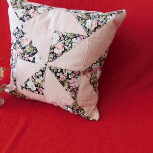 May include: A square throw pillow featuring a patchwork design of solid light pink fabric triangles alternating with black fabric triangles printed with a floral pattern of small pink, white, and green flowers. The pillow has a piped edge.