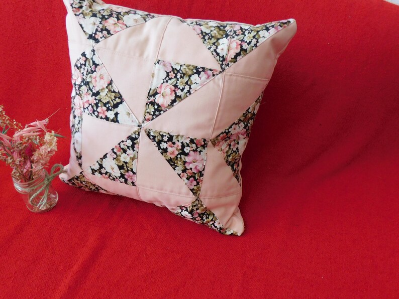 May include: A square throw pillow featuring a patchwork design of solid light pink fabric triangles alternating with black fabric triangles printed with a floral pattern of small pink, white, and green flowers. The pillow has a piped edge.