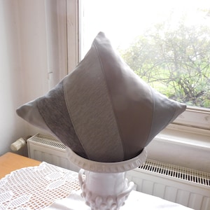 Könnte beinhalten: Ein quadratisches Zierkissen mit einem Patchwork-Design in Grautönen. Das Kissen liegt auf einer weißen Keramikvase. Das Kissen hat eine Vielzahl von Texturen und Materialien, darunter glatte und strukturierte Stoffe.