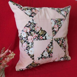 May include: A square throw pillow featuring a patchwork design of solid light pink fabric triangles alternating with black fabric triangles printed with a floral pattern of pink, white, and green flowers. The pillow has a smooth, finished texture.