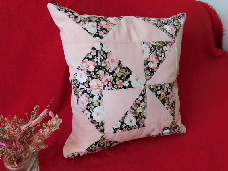 May include: A square throw pillow featuring a patchwork design of solid light pink fabric triangles alternating with black fabric triangles printed with a floral pattern of pink, white, and green flowers. The pillow has a smooth, finished texture.
