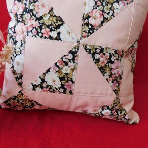 May include: A square throw pillow featuring a patchwork design of solid light pink fabric alternating with a black floral print fabric. The floral pattern consists of pink, white, and green flowers on a black background, arranged in triangles.