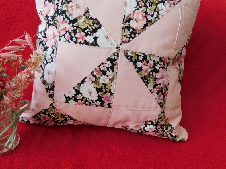 May include: A square throw pillow featuring a patchwork design of solid light pink fabric alternating with a black floral print fabric. The floral pattern consists of pink, white, and green flowers on a black background, arranged in triangles.