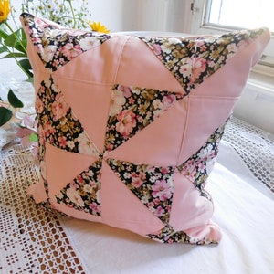 Könnte beinhalten: Ein quadratisches Zierkissen mit einem Patchwork-Design. Das Kissen hat abwechselnde Abschnitte aus einfarbigem hellrosa Stoff und Stoff mit Blumendruck mit rosa, weißen und schwarzen Blumen. Das Kissen liegt auf einer weißen Oberfläche.