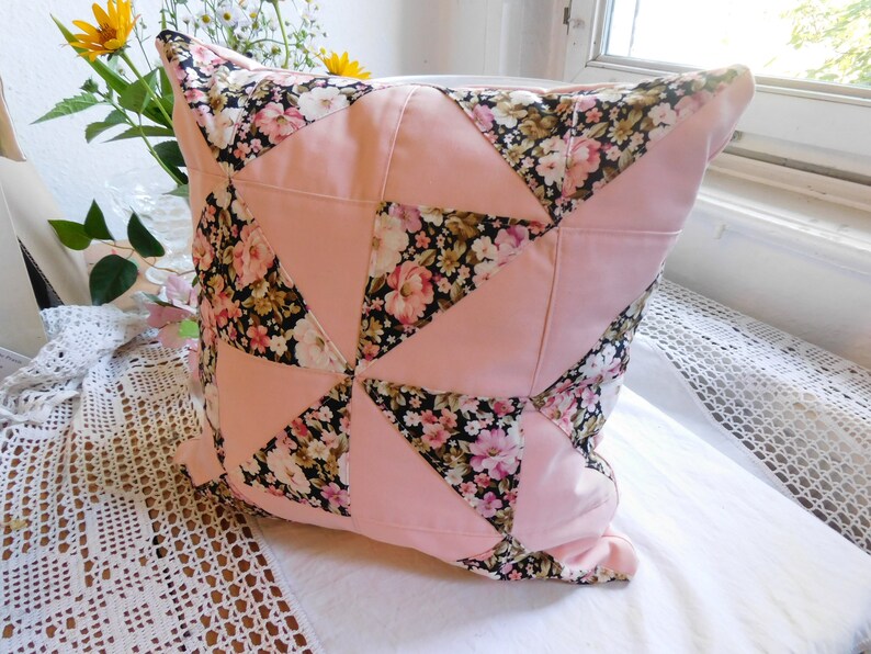 May include: A square throw pillow featuring a patchwork design of alternating solid light pink triangles and black fabric triangles printed with a floral pattern of pink, white, and yellow flowers with green leaves, arranged in a pinwheel pattern.