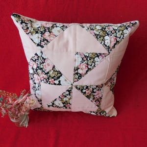 May include: A square throw pillow featuring a patchwork design of solid light pink fabric triangles alternating with black fabric triangles printed with a floral pattern of white, pink, and green flowers. The pillow has a piped edge finish.