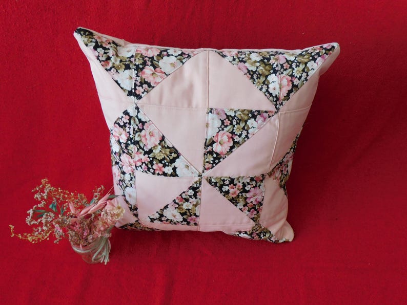 May include: A square throw pillow featuring a patchwork design of solid light pink fabric triangles alternating with black fabric triangles printed with a floral pattern of white, pink, and green flowers. The pillow has a piped edge finish.
