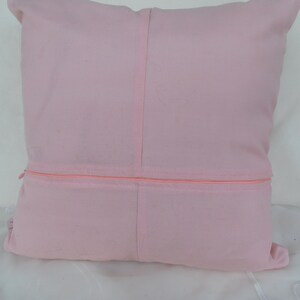 May include: A square light pink throw pillow cover featuring a central vertical seam and a horizontal zipper closure across the middle. The fabric has a smooth, matte texture and a consistent pastel pink color throughout the entire square surface.