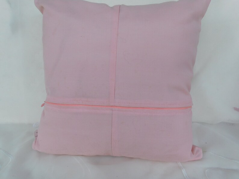 May include: A square light pink throw pillow cover featuring a central vertical seam and a horizontal zipper closure across the middle. The fabric has a smooth, matte texture and a consistent pastel pink color throughout the entire square surface.