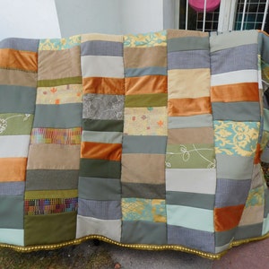 Könnte beinhalten: Ein Patchwork-Quilt mit einer Vielzahl von Stoffen in Grün-, Orange-, Beige- und Grautönen. Der Quilt weist rechteckige Blöcke mit unterschiedlichen Texturen und Mustern auf, darunter florale und geometrische Designs. Die Kanten sind mit einem goldenen Rand versehen.