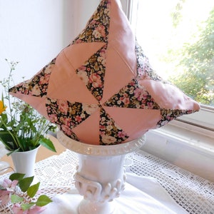 May include: A square throw pillow featuring a patchwork design of solid light pink fabric triangles alternating with black fabric triangles printed with a floral pattern of small pink, white, and yellow flowers and green leaves.
