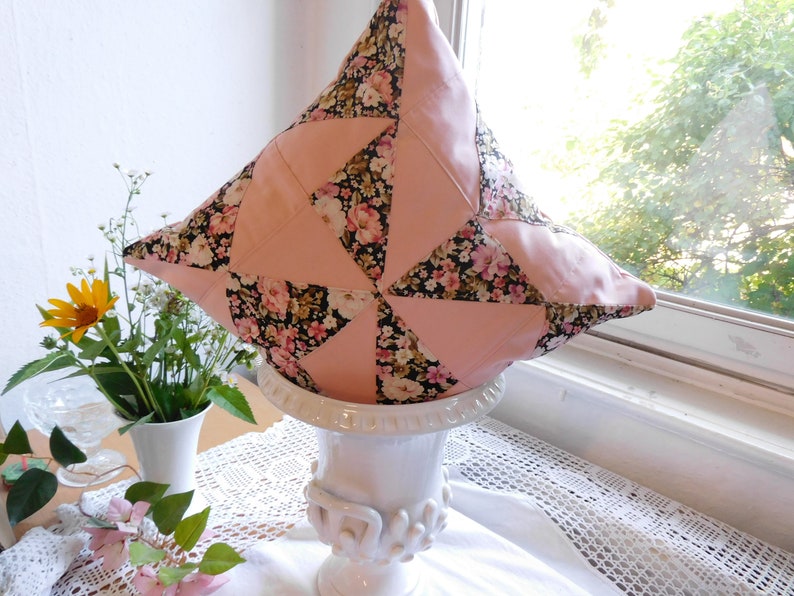 May include: A square throw pillow featuring a patchwork design of solid light pink fabric triangles alternating with black fabric triangles printed with a floral pattern of small pink, white, and yellow flowers and green leaves.