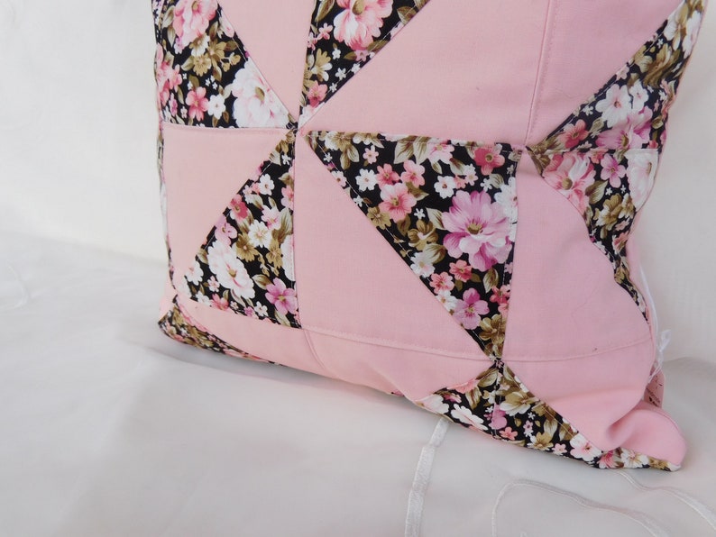 May include: A square throw pillow featuring a patchwork design of solid light pink fabric triangles alternating with black fabric triangles printed with a floral pattern of pink, white, and tan flowers and green leaves, stitched together with visible seams.