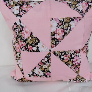 May include: A square throw pillow featuring a patchwork design of solid light pink fabric and a black fabric with a floral pattern of pink, white, and green flowers. The geometric arrangement creates a pinwheel effect across the surface.