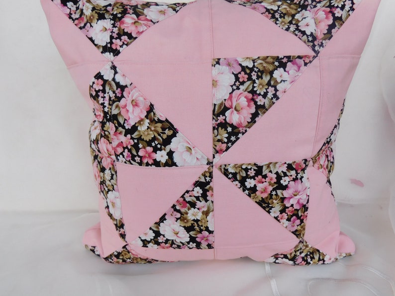 May include: A square throw pillow featuring a patchwork design of solid light pink fabric and a black fabric with a floral pattern of pink, white, and green flowers. The geometric arrangement creates a pinwheel effect across the surface.