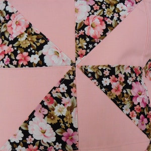 May include: A square throw pillow cover featuring a patchwork design of solid light pink fabric alternating with a black fabric printed with a floral pattern of pink, white, and tan flowers. The pieces are stitched together in a pinwheel pattern.
