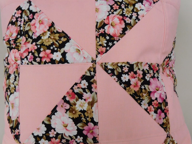 May include: A square throw pillow cover featuring a patchwork design of solid light pink fabric alternating with a black fabric printed with a floral pattern of pink, white, and tan flowers. The pieces are stitched together in a pinwheel pattern.