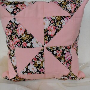May include: A square throw pillow featuring a patchwork design of solid light pink fabric and a black fabric with a multi-colored floral print. The pattern consists of alternating triangles and squares arranged in a geometric pinwheel configuration.