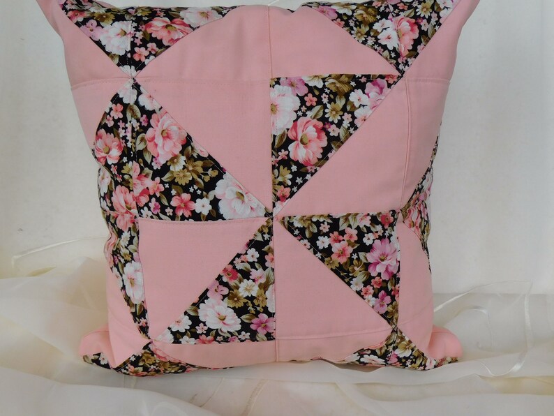 May include: A square throw pillow featuring a patchwork design of solid light pink fabric and a black fabric with a multi-colored floral print. The pattern consists of alternating triangles and squares arranged in a geometric pinwheel configuration.