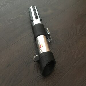 Pu&ograve; includere: Una spada laser argentata con un'impugnatura nera e un pulsante rosso e giallo. La spada laser &egrave; su una superficie di legno scuro.