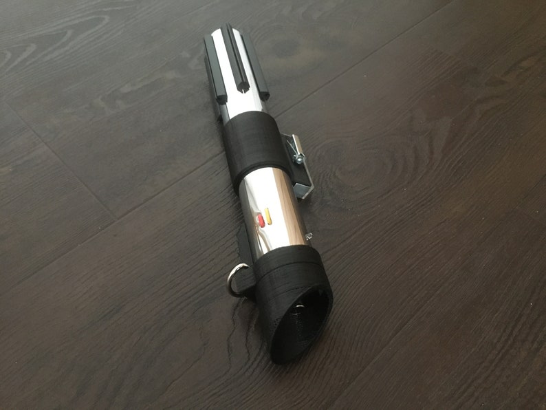 Pu&ograve; includere: Una spada laser argentata con un'impugnatura nera e un pulsante rosso e giallo. La spada laser &egrave; su una superficie di legno scuro.