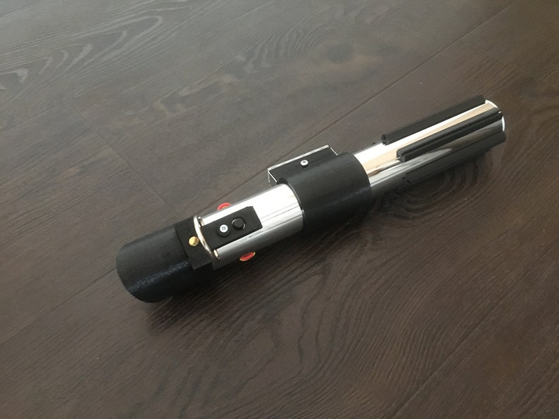 Pu&ograve; includere: Un accessorio per spada laser nero e argento con un'impugnatura nera e una lama argentata. L'impugnatura ha alcuni pulsanti e un piccolo accento dorato.