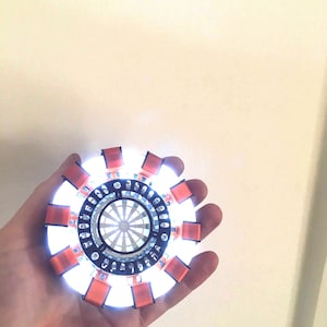 May include: A close-up of a light-up Iron Man Arc Reactor replica. The reactor is made of plastic and has a silver and orange colour scheme. The reactor is lit up with white LEDs.