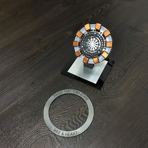 Może przedstawiać: Srebrny metalowy pierścień z wygrawerowanym napisem "PROOF THAT TONY STARK HAS A HEART". Pierścień leży na ciemnym drewnianym tle obok srebrnego i miedzianego modelu reaktora Arc Iron Mana.