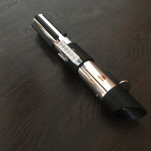 Pu&ograve; includere: Un accessorio per lightsaber argento e nero con un'impugnatura nera e una lama argentata. L'impugnatura ha un'impugnatura nera e un pulsante argentato con un accento rosso e giallo.