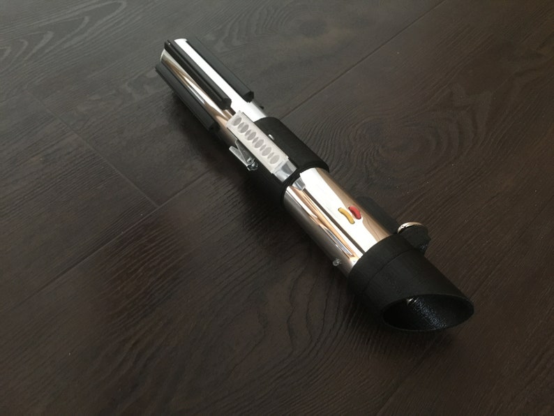 Pu&ograve; includere: Un accessorio per lightsaber argento e nero con un'impugnatura nera e una lama argentata. L'impugnatura ha un'impugnatura nera e un pulsante argentato con un accento rosso e giallo.