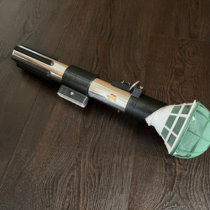Pu&ograve; includere: Una spada laser argentata e nera con una palla di schiuma floreale verde attaccata all'estremit&agrave;. La spada laser &egrave; su un pavimento di legno scuro.