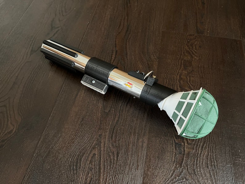 Pu&ograve; includere: Una spada laser argentata e nera con una palla di schiuma floreale verde attaccata all'estremit&agrave;. La spada laser &egrave; su un pavimento di legno scuro.